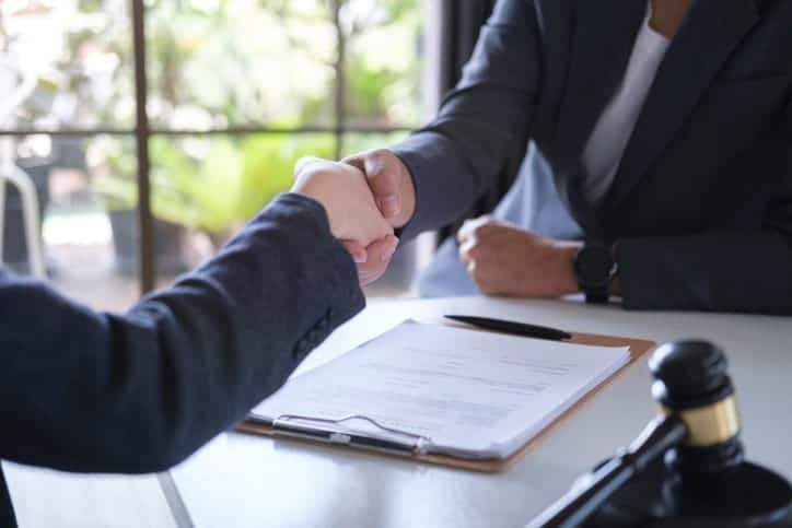 A wrongful death attorney is shaking hands with her client. Between them is paperwork and a gavel. 