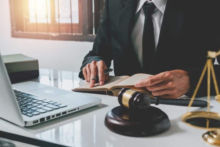 A personal injury lawyer is working in a notebook at his desk. In front of him is an open laptop, a gavel, and the scales of justice. 