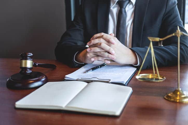 A personal injury attorney sits at his desk with his hands clasped. Around him is a gavel, paperwork, and the scales of justice.